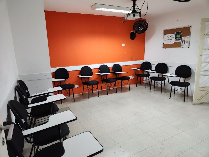 Sala de aula com cadeiras pretas, mesas brancas e parede laranja